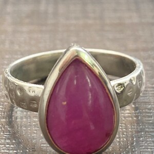 Puede incluir: Un anillo de plata con una piedra preciosa rosa en forma de lágrima engastada en un engaste de plata. La banda del anillo está texturizada con pequeños bultos.