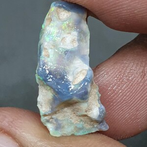 May include: A raw opal gemstone, displaying a mix of colours. The opal has a blue and white base with flashes of green and yellow. The stone is held between two fingers, highlighting its natural, uncut form.