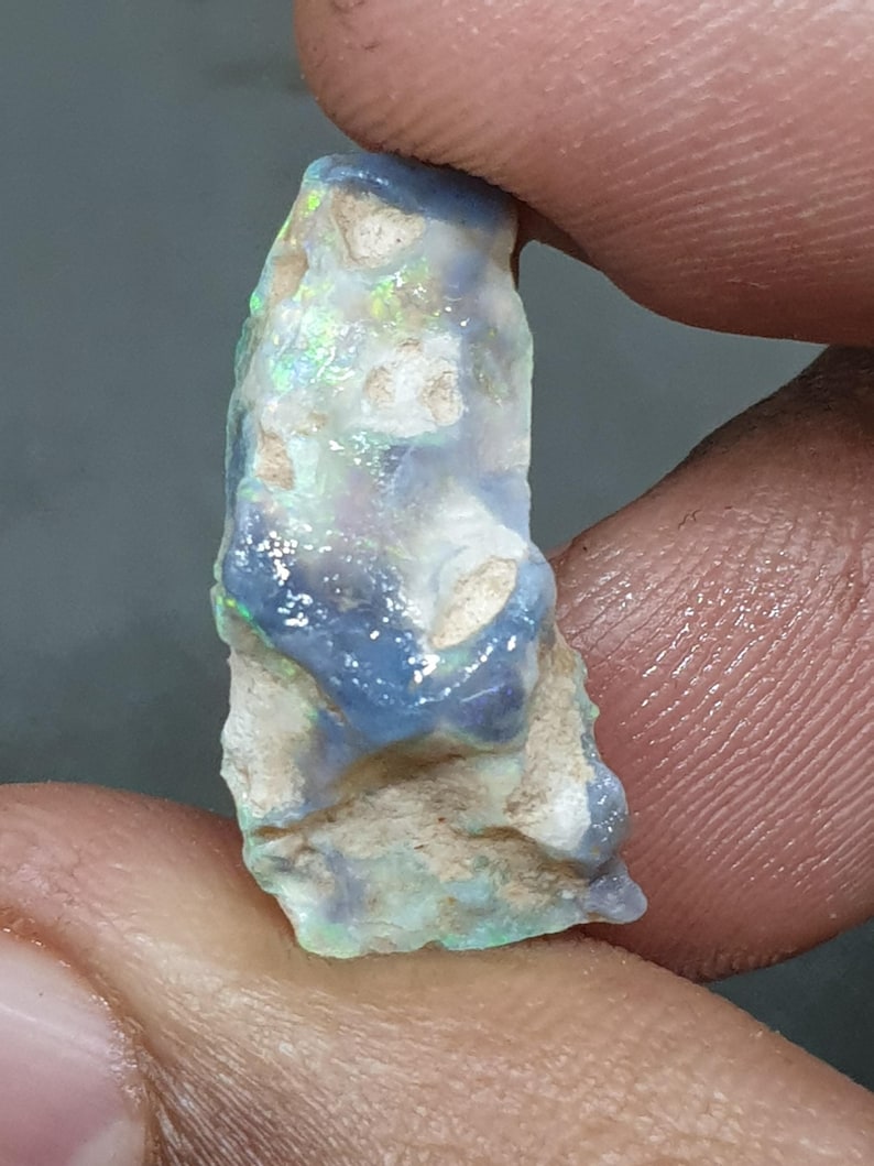 May include: A raw opal gemstone, displaying a mix of colours. The opal has a blue and white base with flashes of green and yellow. The stone is held between two fingers, highlighting its natural, uncut form.