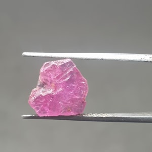 May include: A raw, uncut pink gemstone held by silver tweezers. The gemstone displays a crystalline structure and a vibrant, translucent colour. The background is a solid, neutral grey.