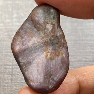May include: A purple and gray rough cut gemstone with a star-like pattern. The gemstone is a natural, unpolished stone.