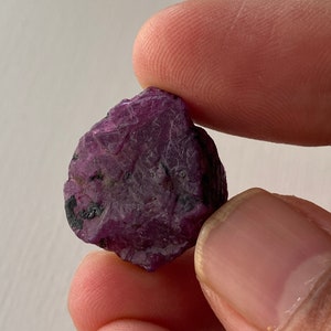 Peut inclure: Une pierre précieuse brute de couleur violette avec une inclusion foncée, presque noire. La pierre précieuse est tenue par une main.