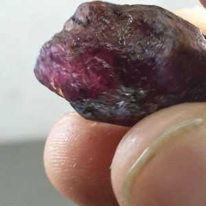 May include: A raw gemstone, predominantly burgundy with purple undertones. The surface is textured, displaying variations in tone. The stone is held between fingers, highlighting its natural, unpolished state. The overall appearance suggests a natural mineral specimen.