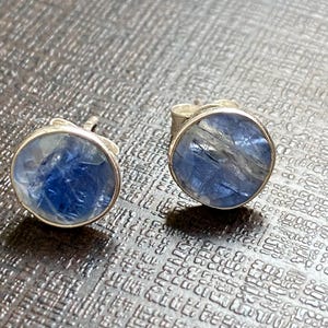 May include: A pair of silver stud earrings with round blue gemstone settings. The gemstones have a shimmery, iridescent quality.