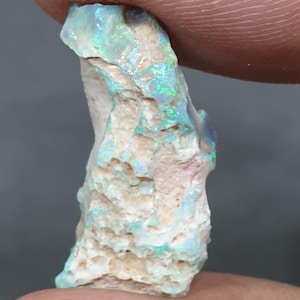 May include: A raw opal gemstone with a light beige base. The opal exhibits flashes of green and blue across its surface. The stone is held between two fingers, highlighting its unique texture and colour variations. The opal is a natural, uncut stone.