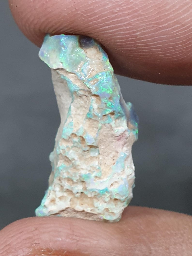 May include: A raw opal gemstone with a light beige base. The opal exhibits flashes of green and blue across its surface. The stone is held between two fingers, highlighting its unique texture and colour variations. The opal is a natural, uncut stone.
