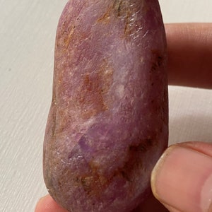 May include: A smooth, oblong, natural stone with a deep reddish-purple hue. The stone has a polished appearance and shows subtle variations in colour and texture, with hints of brown and lighter purple. The stone is held between fingers.