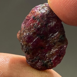 May include: A rough, dark red ruby gemstone with a natural, unpolished finish. The ruby has a deep, rich colour and is a beautiful example of this precious stone.