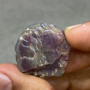May include: A raw, uncut gemstone, held between fingers, displaying a deep purple colour. The stone has a textured surface with visible ridges and colour variations, from dark purple to lighter shades.