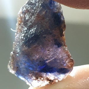 May include: A close-up of a natural, rough-cut blue gemstone. The stone has a textured surface with areas of deep blue and brown hues. The gemstone is held between two fingers, showcasing its irregular shape and natural beauty.
