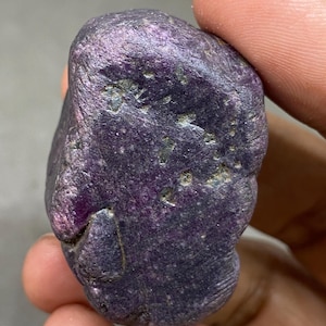 May include: A rough, natural gemstone with a deep purple hue. The surface has a textured appearance with subtle variations in color and small, dark inclusions. The stone is held in a hand, showcasing its irregular shape and natural beauty.