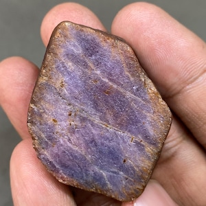 May include: A rough, purple and brown gemstone with a striated surface. The gemstone is held in a hand.
