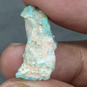 May include: A raw opal gemstone with a mix of colours. The opal displays flashes of green and blue, set against a light-coloured, textured base. The stone is held between fingers, showcasing its raw, unpolished state.