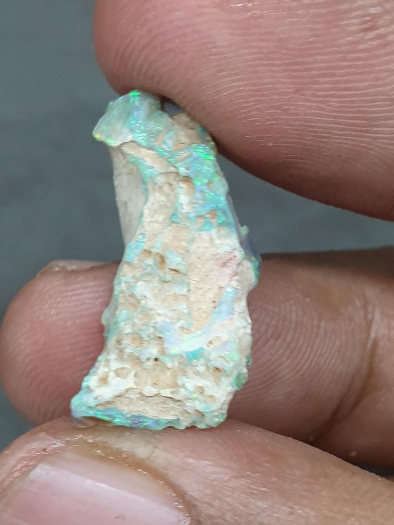 May include: A raw opal gemstone with a mix of colours. The opal displays flashes of green and blue, set against a light-coloured, textured base. The stone is held between fingers, showcasing its raw, unpolished state.