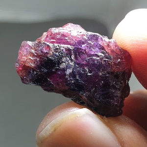 May include: A natural, unpolished gemstone in shades of purple and reddish-pink. The rough texture of the mineral is visible, with light reflecting off its surface. The stone is held between fingers.