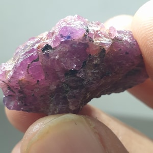 May include: A raw ruby gemstone, displaying a rich pink and purple colour. The natural stone has a textured surface with small, irregular facets. The ruby is held between two fingers, highlighting its raw, unpolished appearance.