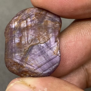 Può includere: Una pietra preziosa grezza di colore viola e marrone con un motivo a forma di stella. La pietra preziosa è tenuta in una mano.