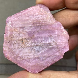 May include: A rough-cut, pink gemstone with a layered, textured appearance. The stone has a soft, light pink hue with subtle variations in color and visible natural lines. The overall shape is irregular, showcasing its raw, unpolished state.