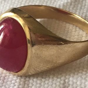 Puede incluir: Un anillo de oro con una gran piedra preciosa de rubí de forma ovalada. El rubí es de color rojo intenso y está engastado en un engaste de bisel.