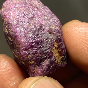 Puede incluir: Una gema de rubí púrpura sin pulir con un ligero tono rojizo. La gema tiene un acabado natural sin pulir.