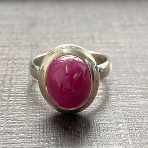 May include: A silver ring with a large, oval, deep pink gemstone. The gemstone is set in a bezel setting, and the ring band is polished silver. The ring is displayed on a textured, grey surface.
