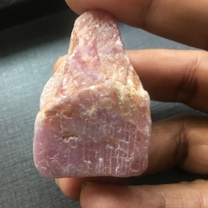 May include: A rough, uncut pink gemstone with a textured surface. The stone is held between fingers, showcasing its natural, irregular shape and color variations. The gem has a crystalline structure.