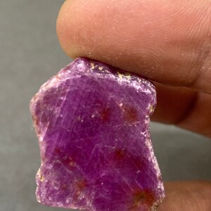 Puede incluir: Una gema de rubí magenta oscuro en bruto. La piedra natural tiene una superficie texturizada con estrías internas visibles y pequeñas inclusiones de color marrón rojizo. El rubí se sostiene entre dos dedos.