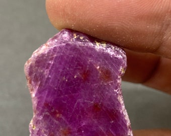 以下が含まれることがあります： 生の濃いマゼンタ色のルビーの宝石。 天然石は、目に見える内部の縞模様と小さな赤褐色のインクルージョンを備えたテクスチャード加工された表面を持っています。 ルビーは2本の指の間に挟まれています。