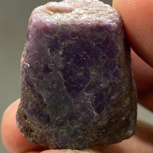 May include: A raw, uncut gemstone with a deep purple and magenta hue. The stone features a rough, crystalline texture with visible natural facets and irregular edges, displaying a mix of opaque and translucent areas throughout its structure.