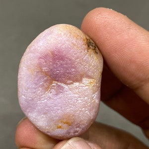Puede incluir: Una piedra preciosa rosa lisa de forma ovalada con un tono púrpura claro e inclusiones marrones.