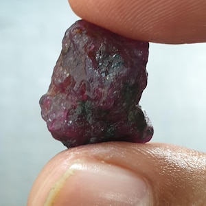 May include: A raw, uncut ruby gemstone held between two fingers. The ruby is a deep, rich red colour with dark inclusions. The surface of the stone appears textured and uneven, showcasing its natural, raw form.