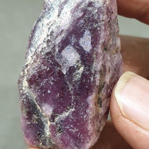 May include: A rough, purple gemstone with a dark grey and white pattern. The gemstone is a raw, uncut ruby.