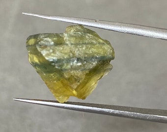 May include: A green and yellow crystal with a rough, faceted surface. The crystal is held in tweezers.