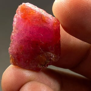 Puede incluir: Una piedra preciosa de rubí rojo intenso con un acabado rugoso y natural. El rubí tiene una forma triangular y una apariencia ligeramente translúcida.
