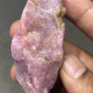 Puede incluir: Un cristal de rubí rosa en bruto con una apariencia translúcida ligeramente nublada. El cristal tiene una forma natural irregular y mide aproximadamente 5 cm de largo.