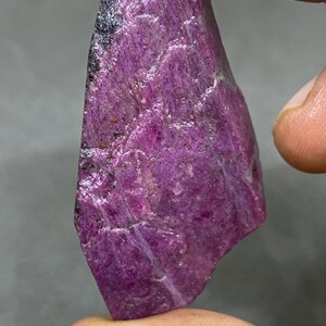 May include: A raw, natural ruby gemstone in a deep magenta colour. The stone has a textured surface with areas of black and lighter pinkish-white veining. The irregular shape is held by a hand, showcasing its raw, unpolished beauty.