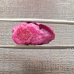 May include: A raw, uncut ruby gemstone, held by tweezers. The ruby is a vibrant pinkish-red colour with a textured surface. The gemstone is irregularly shaped, showcasing its natural, unpolished state. The background is a neutral, textured surface.