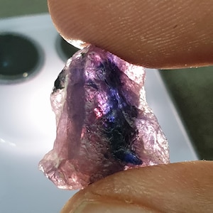 May include: A rough gemstone, held between fingers, displaying a mix of purple, blue, and black colours. The uncut stone has a textured surface, with light reflecting off the facets, creating a shimmering effect.