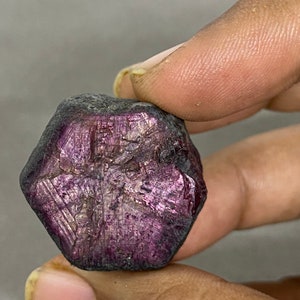 May include: A deep purple, faceted gemstone with a hexagonal shape. The gemstone has a rough, natural texture and is a raw, uncut ruby.