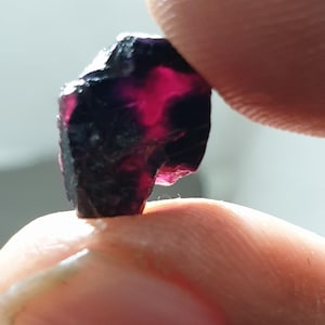 May include: A raw, uncut gemstone with a deep burgundy and black colour. The stone has a rough, textured surface and a translucent, glowing interior. The gemstone is held between two fingers.