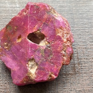 May include: A rough-cut, natural ruby gemstone with a deep pink hue. The stone has a hole in the center and a textured surface. The ruby is a vibrant shade of pink with areas of lighter and darker tones.