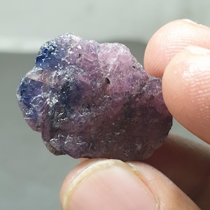 May include: A raw gemstone, displaying a blend of purple and blue shades. The natural mineral has a textured surface. The stone is held between fingers. The colour shifts from deep blue to a lighter purple.