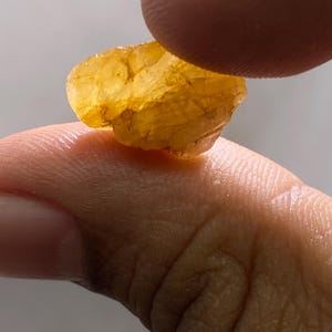 May include: A translucent, golden-yellow gemstone held between two fingers. The rough-cut stone has a textured surface and irregular shape, catching the light. The gemstone appears to be a natural mineral specimen.