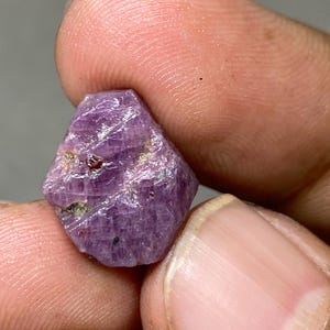 Puede incluir: Una piedra preciosa de rubí de color púrpura intenso con un corte facetado. El rubí tiene una textura rugosa y natural y es de un color púrpura intenso y rico.