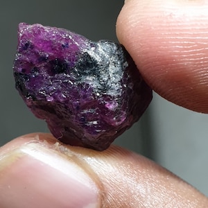 May include: A rough, uncut gemstone in a deep purple colour, with areas of black. The stone's rough surface is visible, showing its natural, unpolished state. The gemstone is held between two fingers, showing its size and shape.