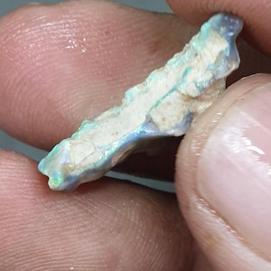 May include: A rough opal specimen, elongated with a white base and iridescent blue, green, and yellow flashes. The opal is held between fingers, showing its natural, unpolished texture and colour variations. The stone has a fractured appearance.