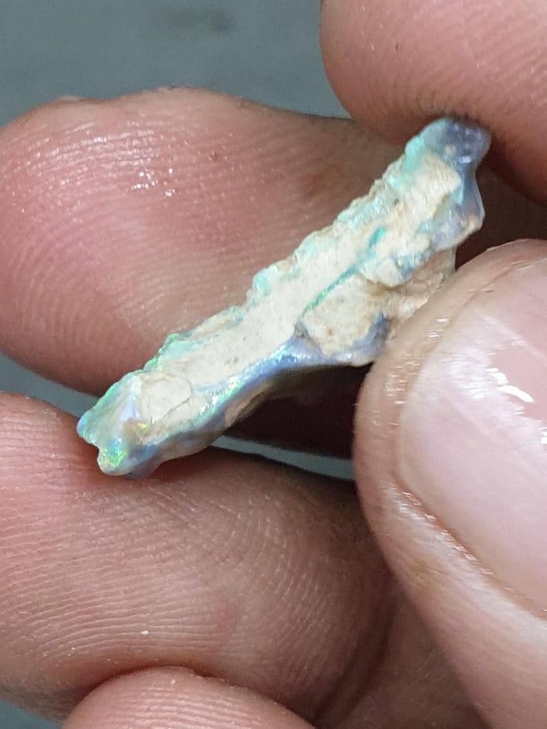 May include: A rough opal specimen, elongated with a white base and iridescent blue, green, and yellow flashes. The opal is held between fingers, showing its natural, unpolished texture and colour variations. The stone has a fractured appearance.