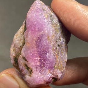Op de afbeelding: Een ruwe, ongeslepen edelsteen met een doorschijnende roze en paarse tint. De steen heeft een ruwe, onregelmatige oppervlaktestructuur met zichtbare kristallijne structuren en kleine vlekjes van donkere, aardse minerale insluitingen in het gesteente.