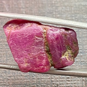 Puede incluir: Una piedra preciosa de rubí rosa en bruto con una forma ligeramente irregular. El rubí se mantiene en su lugar por unas pinzas.