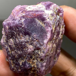 May include: A rough, purple and grey gemstone with a striated surface. The gemstone is a raw, uncut ruby.